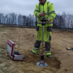 Pracownik w odblaskowym ubraniu obsługuje urządzenie do badania gruntu w piaszczystym wykopie, obok otwarty walizkowy miernik.