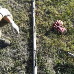 Metalowa rura z otworami, prawdopodobnie wiertnicza, leży na trawie obok rękawic roboczych i fragmentu buta. Na rurze widoczne ślady ziemi.