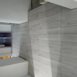 Ściana wyłożona jasnymi, podłużnymi płytkami imitującymi drewno, widoczne półki z wyposażeniem w tle.