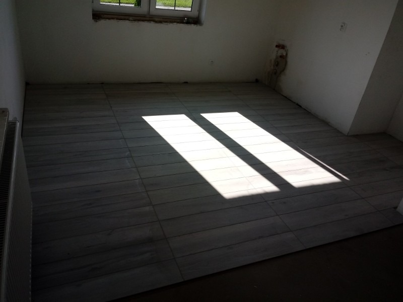 Ułożone szare płytki podłogowe imitujące drewno, z widocznym promieniem słonecznym wpadającym przez okno, ściany w stanie surowym, widoczny grzejnik.