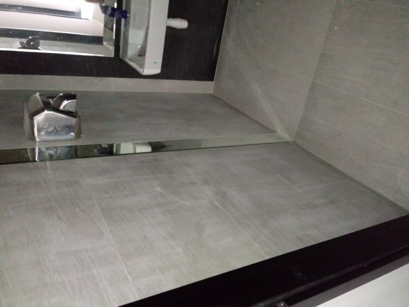 Szare płytki podłogowe i ścienne w łazience, z widocznym chromowanym wykończeniem listwy przypodłogowej i dozownikiem mydła w tle, oraz fragment umywalki z baterią.