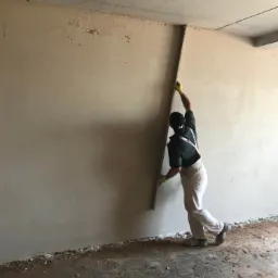 Pracownik w białych spodniach i żółtych rękawicach wygładza świeżo nałożony tynk na ścianie za pomocą długiej łaty. Widoczny fragment sufitu i podłogi z resztkami zaprawy.