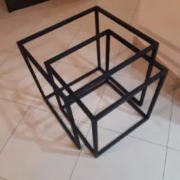 Zestaw trzech metalowych, czarnych, geometrycznych stolików kawowych w kształcie sześcianów o różnych rozmiarach, umieszczonych jeden w drugim na jasnej, kafelkowej podłodze, w tle ściana.