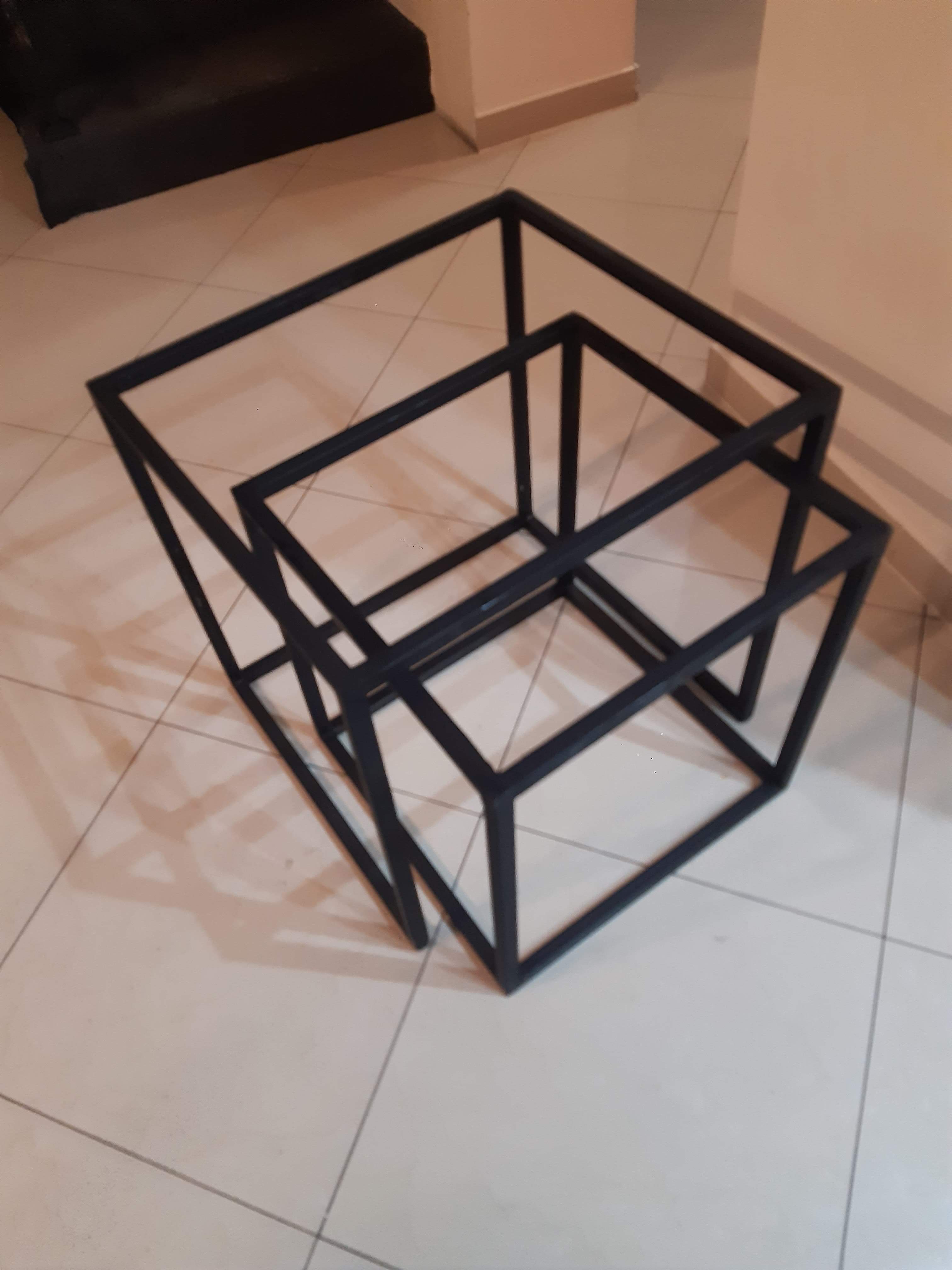 Zestaw trzech metalowych, czarnych, geometrycznych stolików kawowych w kształcie sześcianów o różnych rozmiarach, umieszczonych jeden w drugim na jasnej, kafelkowej podłodze, w tle ściana.