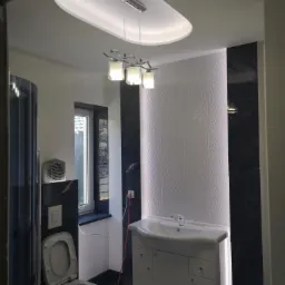 Wykończona łazienka z białą armaturą, kabiną prysznicową, podwieszanym sufitem z oświetleniem LED i czarno-białymi płytkami imitującymi marmur na ścianach.