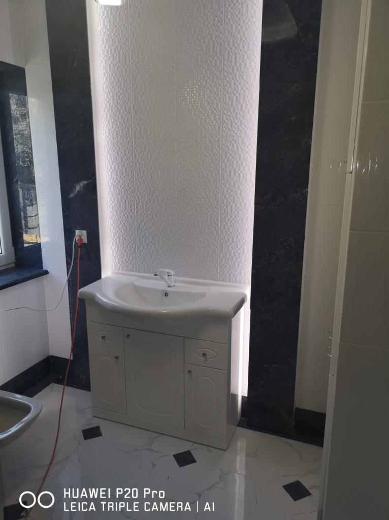 Wykończona łazienka z białą umywalką na szafce, mozaiką na ścianie i czarnymi płytkami imitującymi marmur. Widoczny fragment podłogi z białych płyt z czarnymi wstawkami.