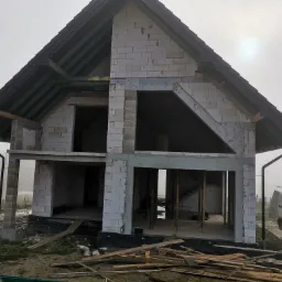 Surowy stan domu jednorodzinnego w trakcie budowy, widoczne ściany z bloczków, więźba dachowa, rynny, w tle mgła, obok zaparkowany biały samochód dostawczy z drabiną na dachu, na pierwszym planie...