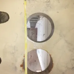 Wykonane dwa otwory w mokrej, betonowej posadzce, widoczny fragment buta roboczego i żółta miara zwijana przy otworach.