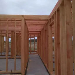 Konstrukcja szkieletowa domu w trakcie budowy, widok wnętrza z betonową podłogą i drewnianym szkieletem ścian, młotek leżący na podłodze.