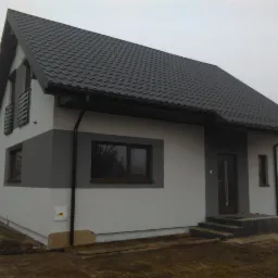 Nowoczesny dom jednorodzinny z dwukolorową elewacją (szary pas na białym tle) i ciemną, metalową dachówką, widok z zewnątrz w pochmurny dzień, z widocznym przyłączem gazu.