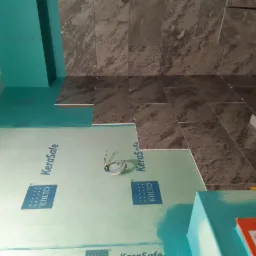 Przygotowanie do układania płytek w łazience: widoczne zabezpieczenie ścian płytami KeraSafe, częściowo ułożone płytki podłogowe w odcieniach szarości, oraz wystające przewody elektryczne.