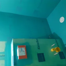 Przygotowanie do prac wykończeniowych w łazience: ściany i sufit pokryte turkusową hydroizolacją KeraSafe, widoczne narożniki, odpływ liniowy i elementy instalacji wodnej. Na ścianie przyklejony...