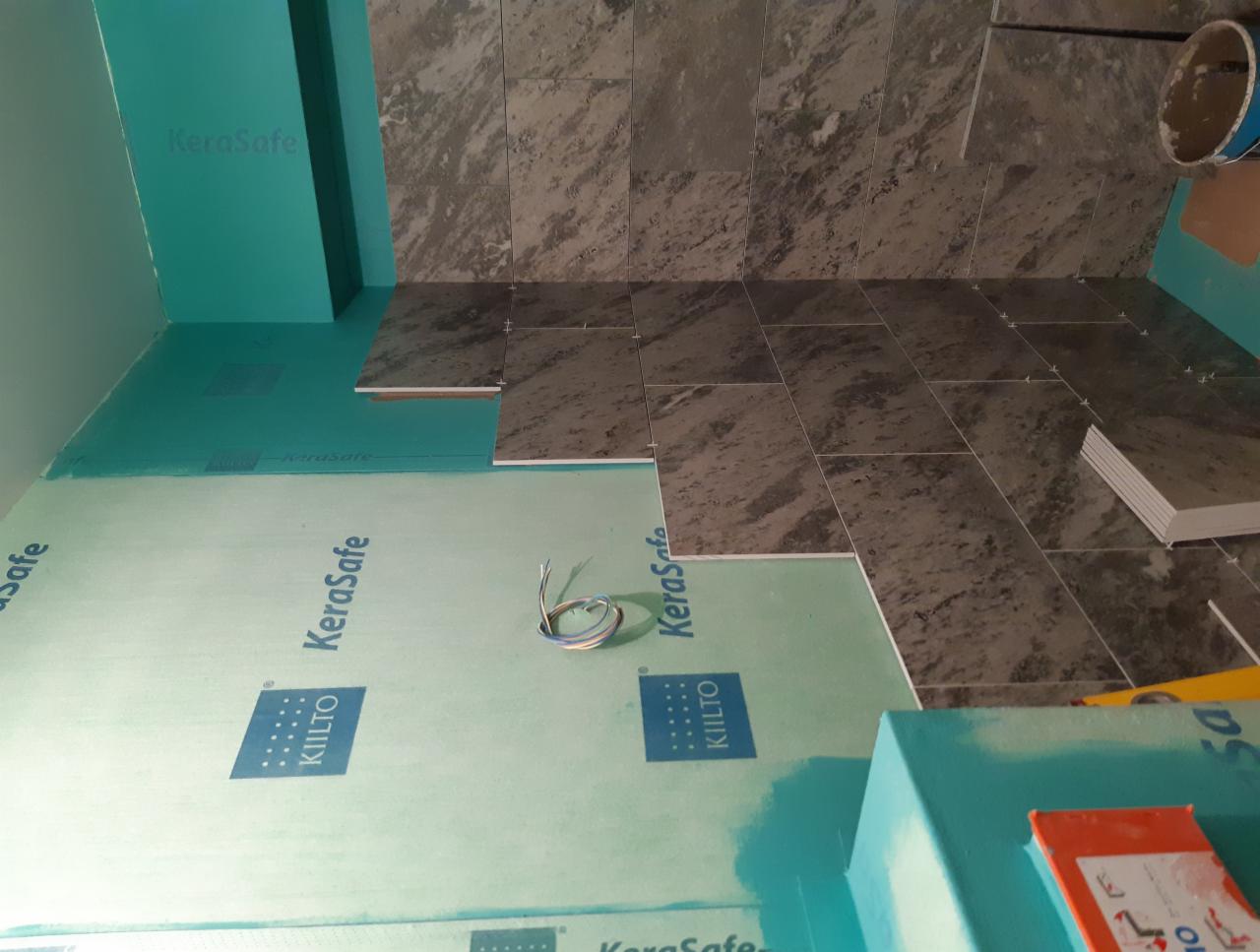 Przygotowanie do układania płytek w łazience: widoczne zabezpieczenie ścian płytami KeraSafe, częściowo ułożone płytki podłogowe w odcieniach szarości, oraz wystające przewody elektryczne.