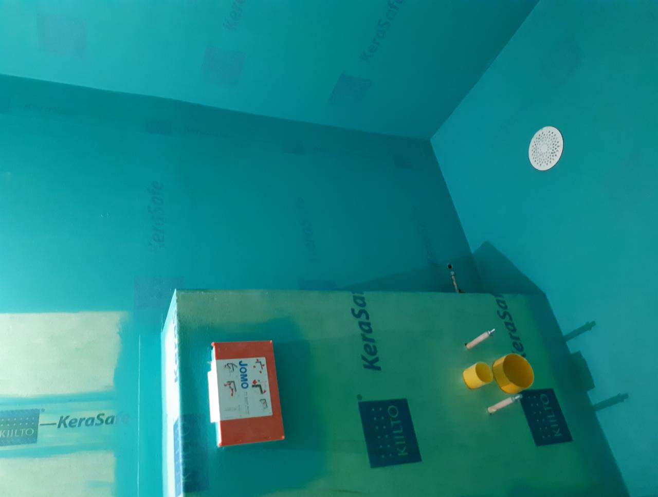 Przygotowanie do prac wykończeniowych w łazience: ściany i sufit pokryte turkusową hydroizolacją KeraSafe, widoczne narożniki, odpływ liniowy i elementy instalacji wodnej. Na ścianie przyklejony...