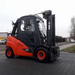 Nowy, pomarańczowo-czarny wózek widłowy marki Linde-Fenwick, sfotografowany z niskiego kąta na tle kostki brukowej i budynków w pochmurny dzień.