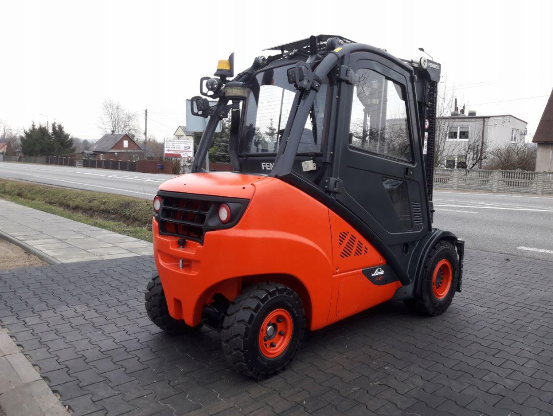 Pomarańczowo-czarny wózek widłowy marki Linde, model H25, z kabiną, stojący na kostce brukowej w pobliżu drogi i zabudowań.