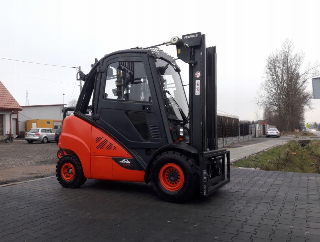 Nowy, pomarańczowo-czarny wózek widłowy marki Linde-Fenwick, sfotografowany z niskiego kąta na tle kostki brukowej i budynków w pochmurny dzień.