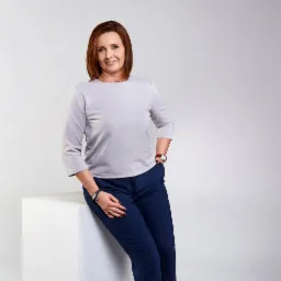 Portret uśmiechniętej kobiety w szarej bluzce i granatowych spodniach, opierającej się o biały sześcian, z zegarkiem na lewym nadgarstku i pierścionkiem na palcu prawej dłoni.