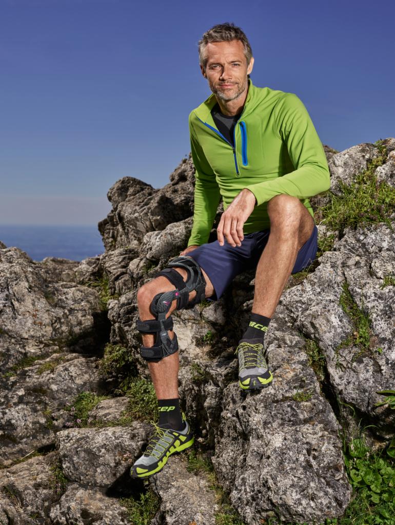 Mężczyzna siedzi na skałach z ortezą na kolanie, ubrany w sportowy strój i buty, w tle błękitne niebo i widok na oddalony krajobraz.