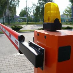 MiKSA - Instalacje Teletechniczne - Automatyczna bariera drogowa z pomarańczową obudową i ostrzegawczym światłem na górze, widoczna na tle parkingu i budynków.