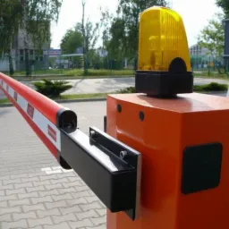 Automatyczna bariera drogowa z pomarańczową obudową i ostrzegawczym światłem na górze, widoczna na tle parkingu i budynków.