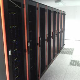 MiKSA - Instalacje Teletechniczne - Rząd szaf serwerowych z pomarańczowymi akcentami w serwerowni, widoczne diody LED i panele kontrolne, biała podłoga z perforowanymi płytami.