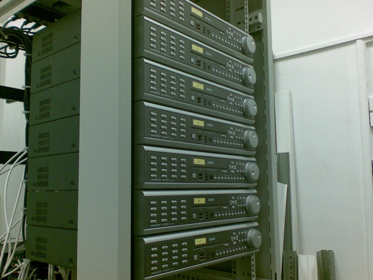 Rząd szarych rejestratorów w szafie rack, widoczne kable i elementy montażowe, oświetlenie sufitowe.