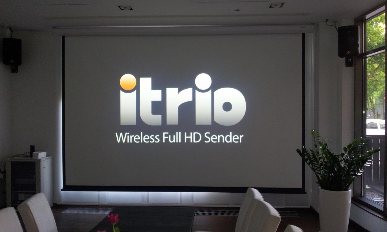 Sala konferencyjna z rozwijanym ekranem projekcyjnym wyświetlającym logo 'itrio Wireless Full HD Sender', głośniki wiszące na ścianach, roślina doniczkowa przy oknie.