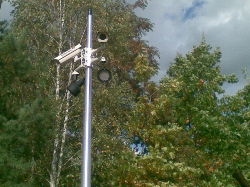 Słup z zamontowanymi kamerami monitoringu i reflektorem, widoczne drzewa w tle.