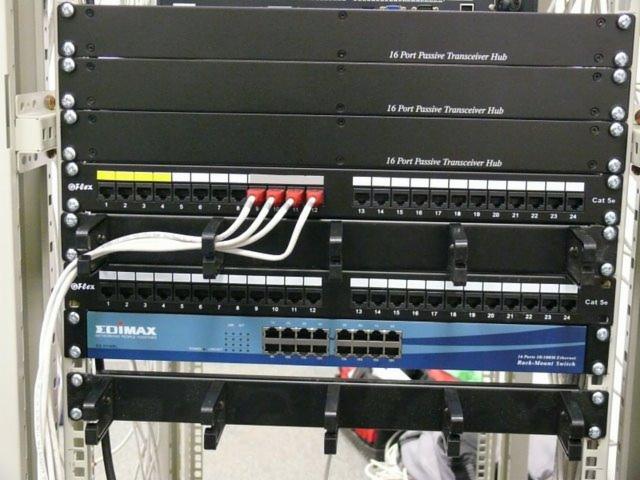 Szafa rack z panelami krosowniczymi, patch panelem z portami RJ45 oznaczonymi numerami, oraz switchem Edimax z niebieskim paskiem, podłączonymi kablami sieciowymi.