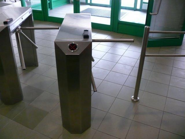 Metalowe bramki obrotowe z czytnikiem kart w budynku z zielonymi drzwiami wejściowymi i szarą podłogą z płytek.