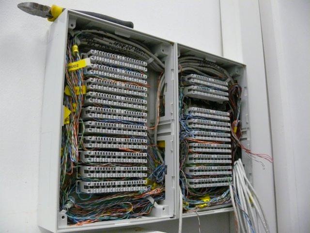 Otwarta skrzynka rozdzielcza z gęsto upakowanymi kablami różnego koloru i złączami, widoczne żółte etykiety i czarne szczypce na górze obudowy.