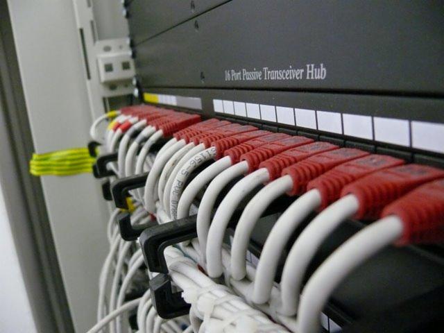 Panel krosowniczy z podłączonymi kablami sieciowymi zakończonymi czerwonymi wtykami RJ45, widoczne białe kable opisane numerami, w tle urządzenie 16 Port Passive Transceiver Hub.