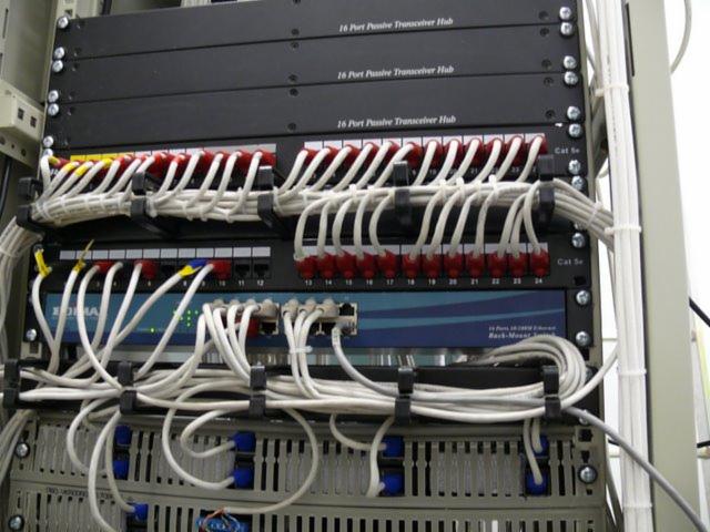 Panel krosowniczy z uporządkowanymi kablami sieciowymi, widoczne porty oznaczone kolorami, czarne uchwyty kablowe utrzymujące porządek, etykiety na urządzeniach.