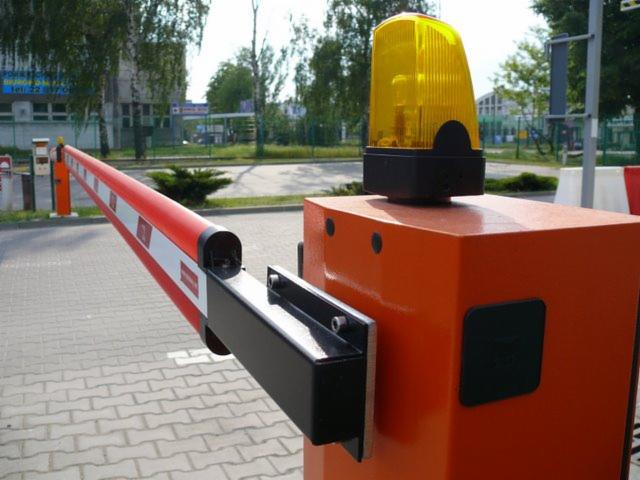 Automatyczna bariera drogowa z pomarańczową obudową i ostrzegawczym światłem na górze, widoczna na tle parkingu i budynków.