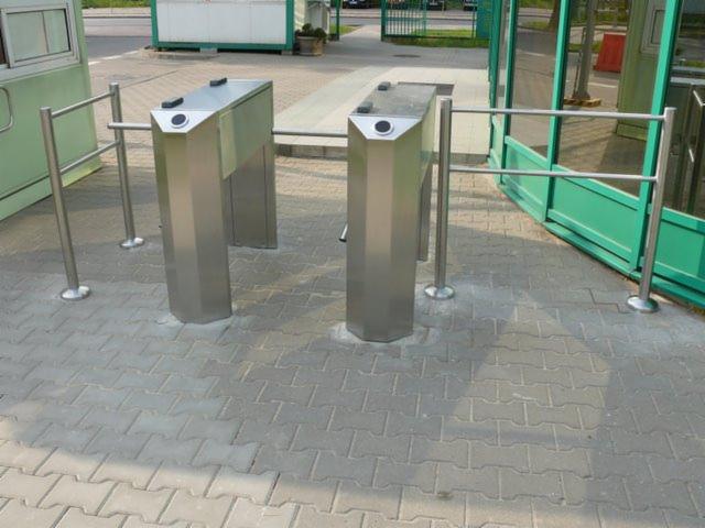 Dwa metalowe, automatyczne kołowroty wejściowe z czytnikami kart, ustawione na szarym bruku przed budynkiem z zielonymi elementami.