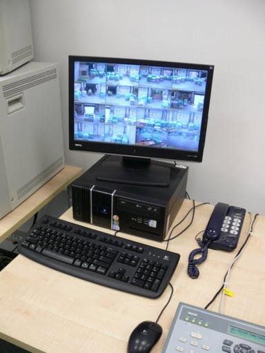 Monitor wyświetlający obraz z wielu kamer monitoringu, komputer stacjonarny, klawiatura, mysz i telefon na biurku w pomieszczeniu biurowym.