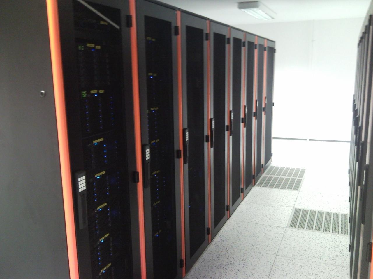 Rząd szaf serwerowych z pomarańczowymi akcentami w serwerowni, widoczne diody LED i panele kontrolne, biała podłoga z perforowanymi płytami.