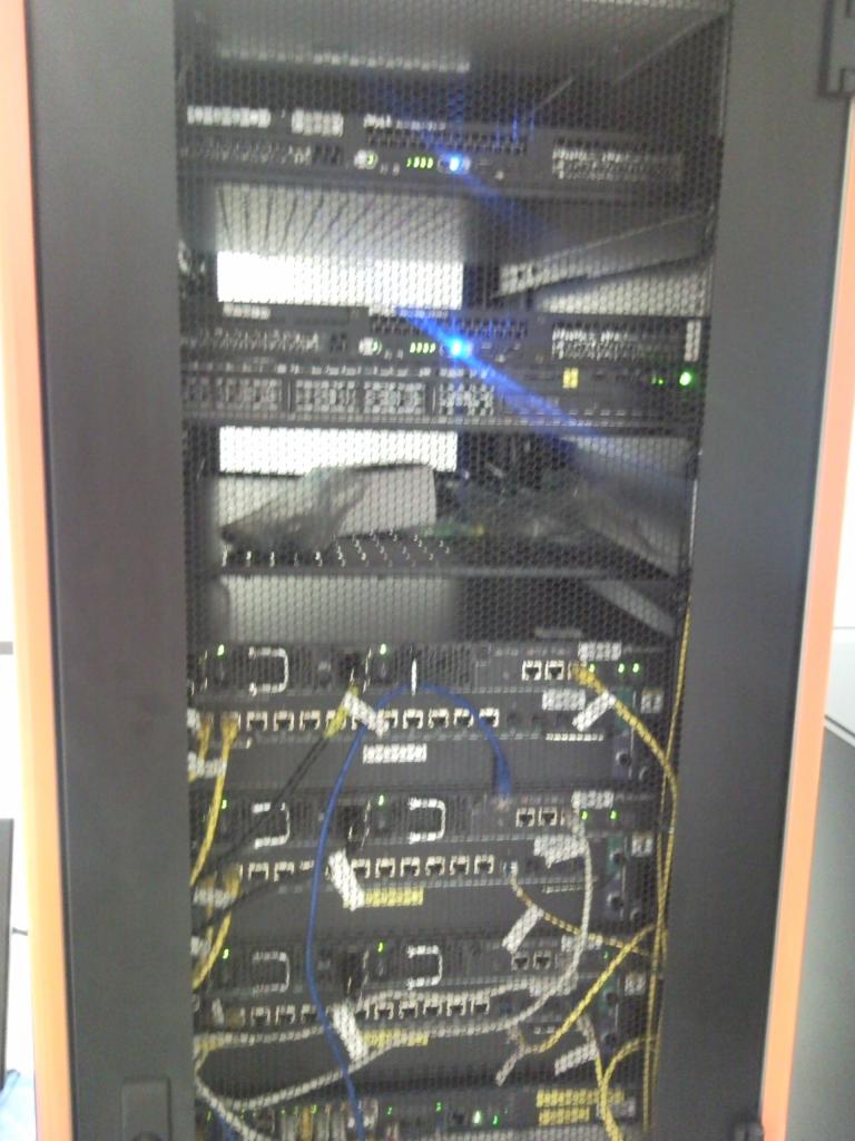 Widok z bliska szafy serwerowej z widocznymi kablami Ethernet, przełącznikami sieciowymi i migającymi diodami LED, ukazujący stan pracy urządzeń.