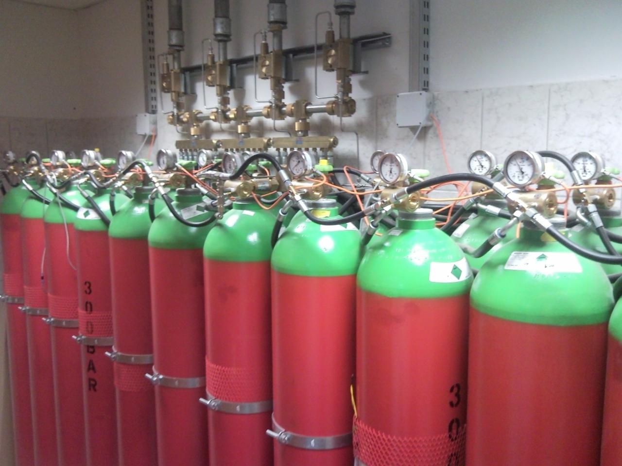 Rząd czerwono-zielonych butli z gazem podłączonych do instalacji z manometrami w pomieszczeniu technicznym.