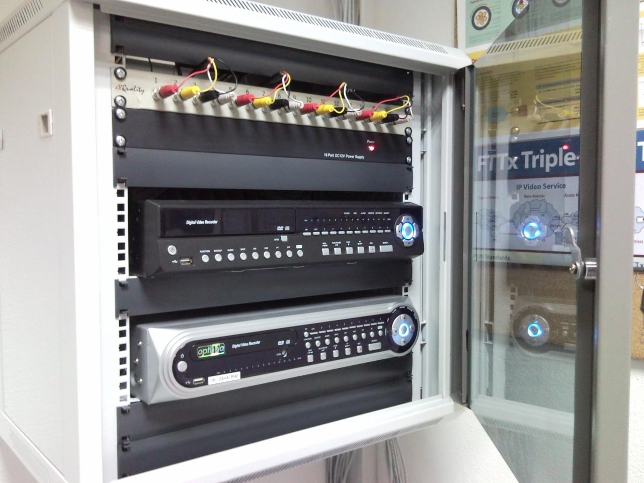 Szafa rack z rejestratorami cyfrowymi i zasilaczem 16-portowym DC12V, zainstalowana w Warszawie, prawdopodobnie do monitoringu IP. Widoczna etykieta 'FTTx Triple IP Video Service' na drzwiach szafy.