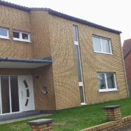 Dwukondygnacyjny dom z żółtej cegły z białymi ramami okiennymi i drzwiami wejściowymi z elementami szklanymi, widoczny trawnik i fragment sąsiedniego budynku z czerwonej cegły.