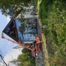 Montaż przeszklonej ściany domu z użyciem podnośnika koszowego, widoczne rusztowania i otoczenie z zielenią.