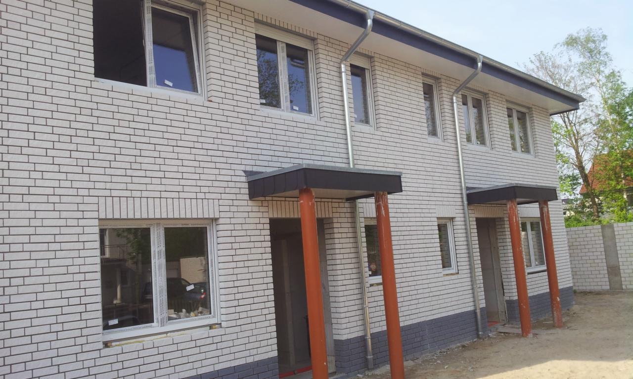 Nowe bliźniacze domy z białej cegły z zamontowanymi oknami, jedno okno otwarte, widoczne rynny i proste zadaszenia nad wejściami wsparte na pomarańczowych kolumnach.