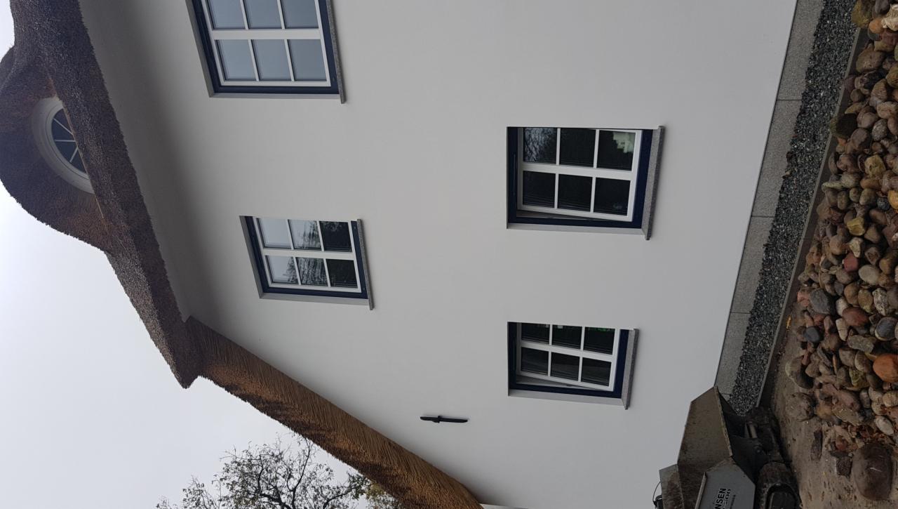 Dom z białymi ścianami i strzechą, widoczne okna z białymi ramami i ciemnymi obwódkami, jedno z okien uchylone, obok domu kamienny chodnik.