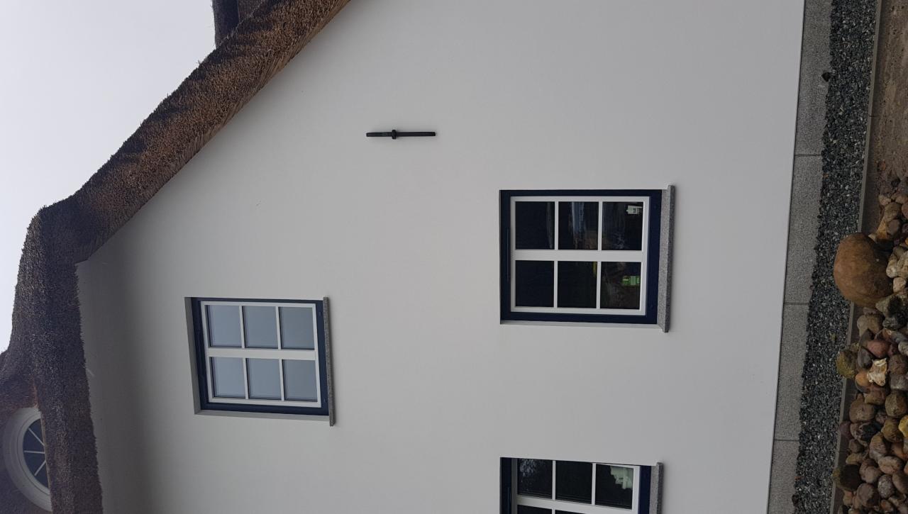 Biała ściana domu krytego strzechą z trzema oknami PCV z ciemnoniebieską ramą i białymi szprosami, widok z boku.