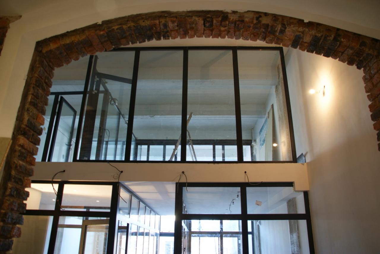 Wnętrze z przeszkloną antresolą z czarnymi ramami, widoczne przez ceglany łuk, drabina oparta o przeszkloną ścianę na górnym poziomie.