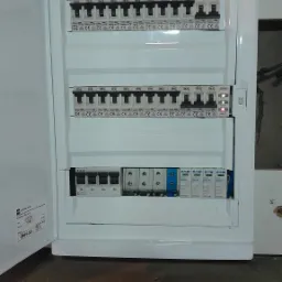 Otwarta biała skrzynka rozdzielcza z bezpiecznikami i wyłącznikami różnicowoprądowymi, widoczne oznaczenia producentów Ideal i Eaton, na podłodze leży czarno-żółty śrubokręt.