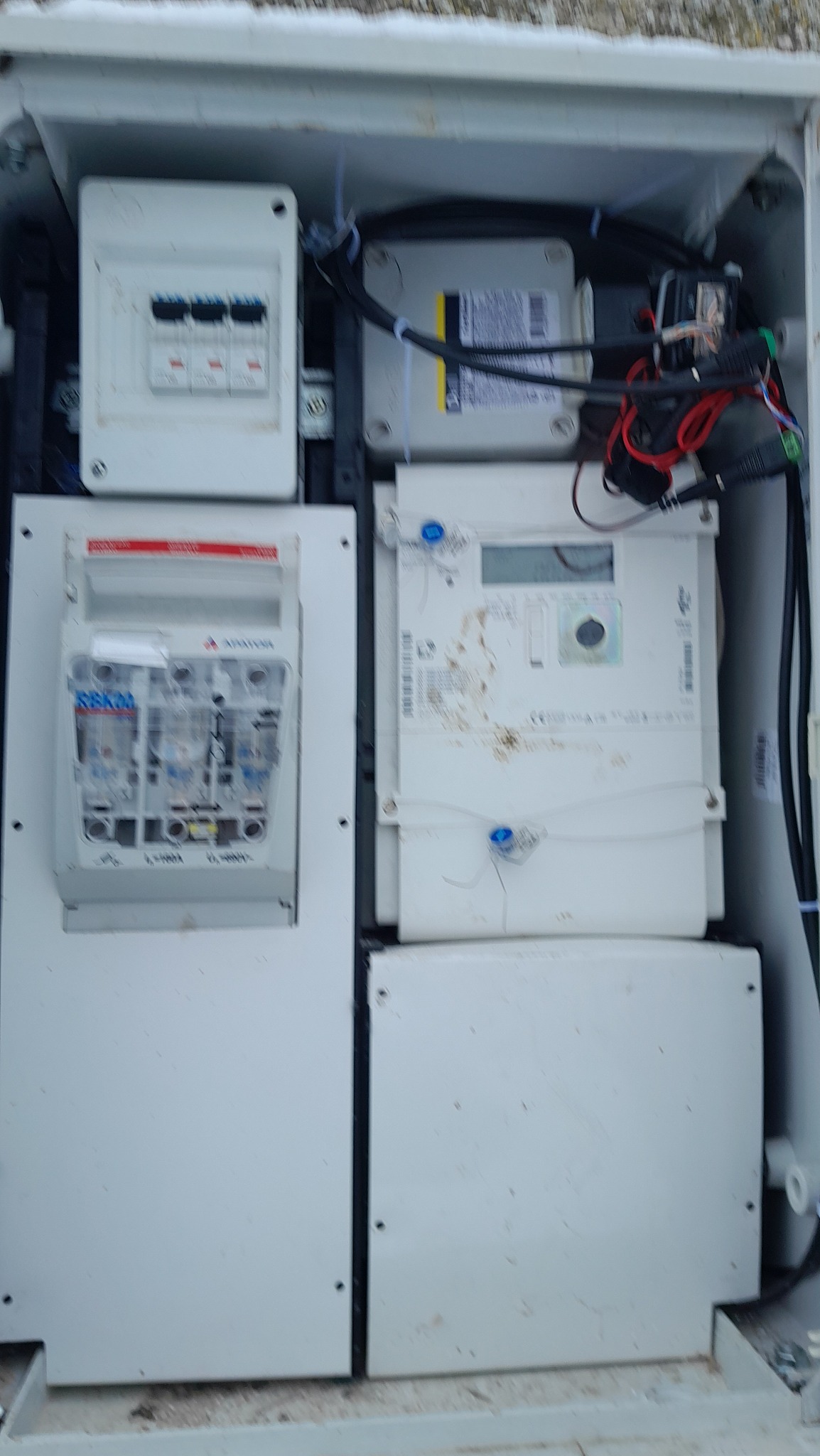 Wnętrze skrzynki elektrycznej z licznikiem, bezpiecznikami i okablowaniem, widoczne ślady użytkowania i zabrudzenia.