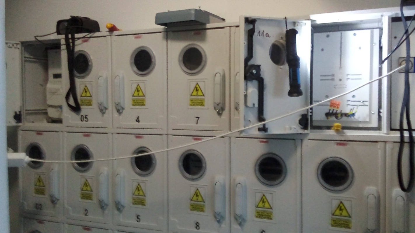 Rząd białych szaf elektrycznych z okrągłymi otworami inspekcyjnymi i ostrzeżeniami o napięciu, jedna szafa otwarta, ukazująca wewnętrzne okablowanie i osprzęt